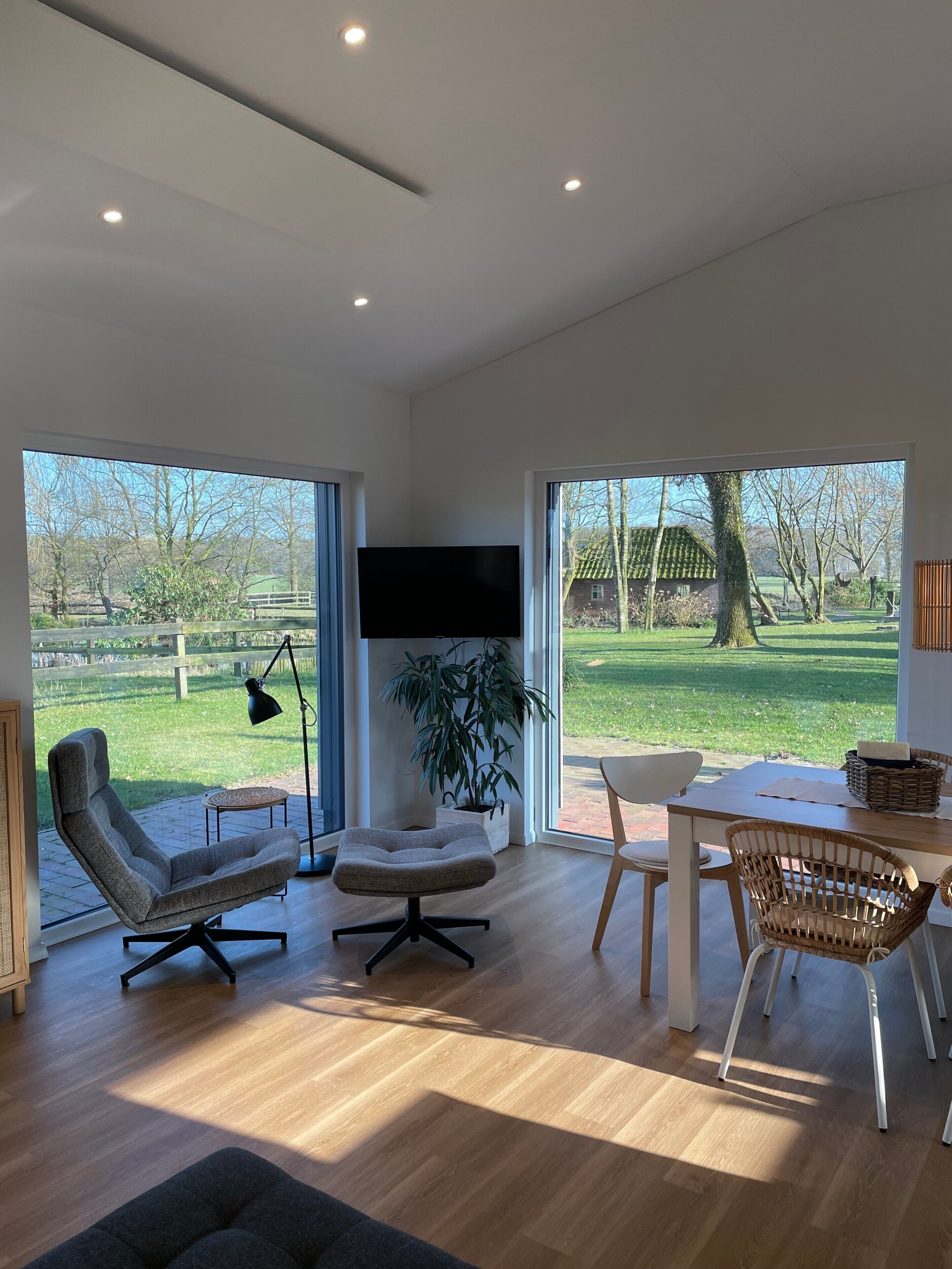 A modern living room with a large TV, comfortable seating, a dining area, and a view of a green landscape.