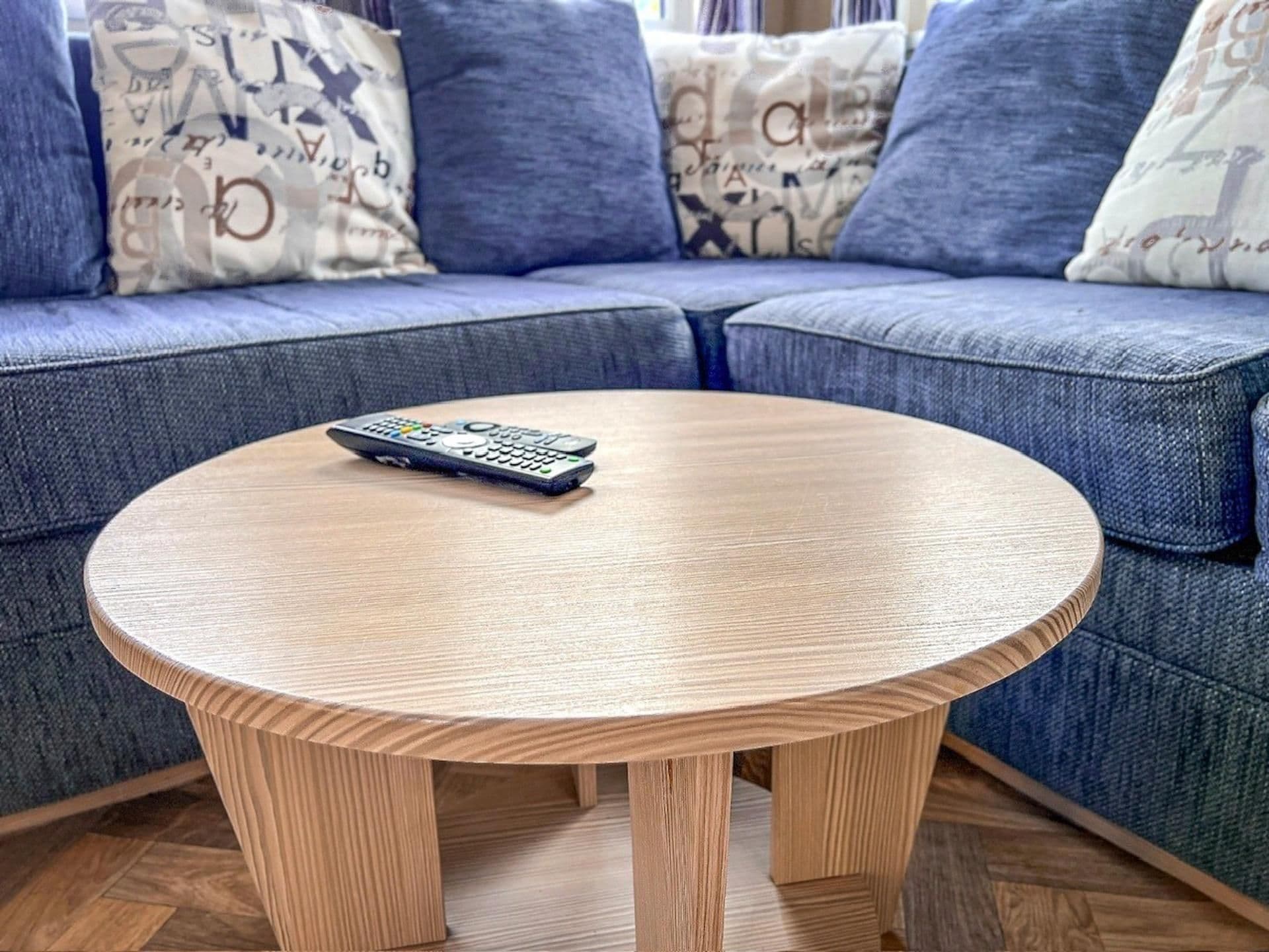A round wooden coffee table with a remote control on it, placed in front of a blue sofa with patterned cushions.
