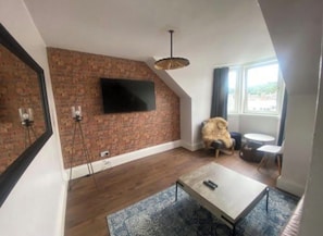 Ein modernes Wohnzimmer mit einer Ziegelwand, einem flachbildfernseher an der Wand über einem Holzboden, einem runden Couchtisch und einem großen Fenster, das für natürliches Licht sorgt.