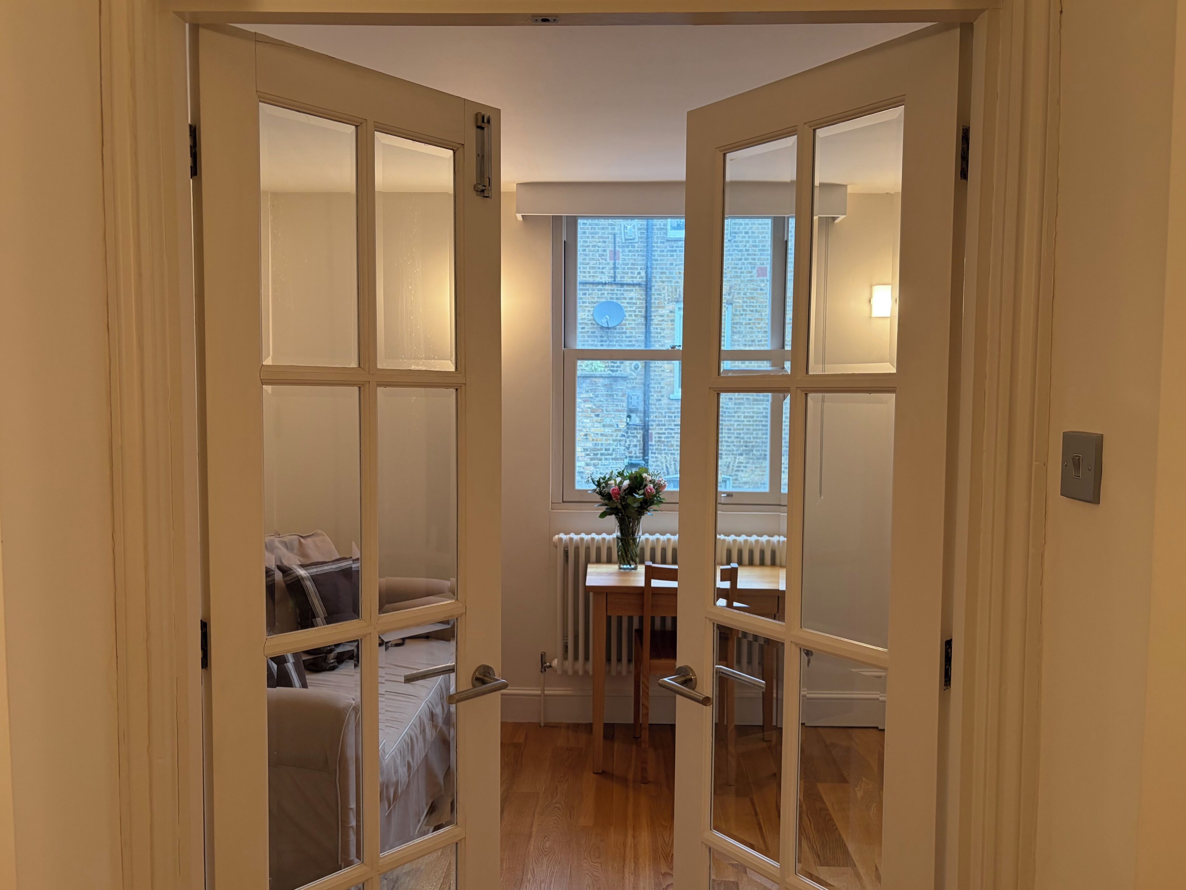 Une pièce avec un plancher en bois, un radiateur, une fenêtre donnant sur un immeuble en briques et un lit recouvert d’une couverture à rayures.
