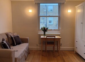 Ein Zimmer mit einem beigen Sofa, einem Holztisch mit Blumen in einer Vase, einem Fenster mit Blick auf ein Backsteinhaus und einem weißen Kleiderschrank.