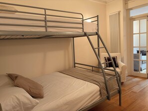 Un lit superposé avec une échelle, un lit simple avec un oreiller, une chaise blanche et une vue sur une autre pièce à travers une porte vitrée.