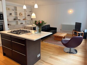 Eine moderne Küche mit Kochinsel, eine Essecke mit schwarzem Sofa und ein Wohnzimmer mit Fernseher.