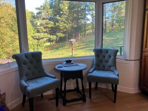 A room with two upholstered chairs and a small round table, a window with a view of a forest, and a wooden cabinet.