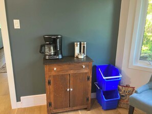 Habitació amb una cafetera, dos recipients d'acer inoxidable, un cub d'emmagatzematge blau i una cadira.
