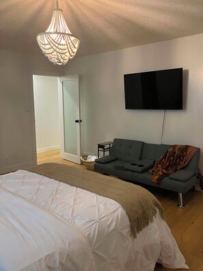 A bedroom with a bed, a grey sofa, a television mounted on the wall, and a door leading to another room.