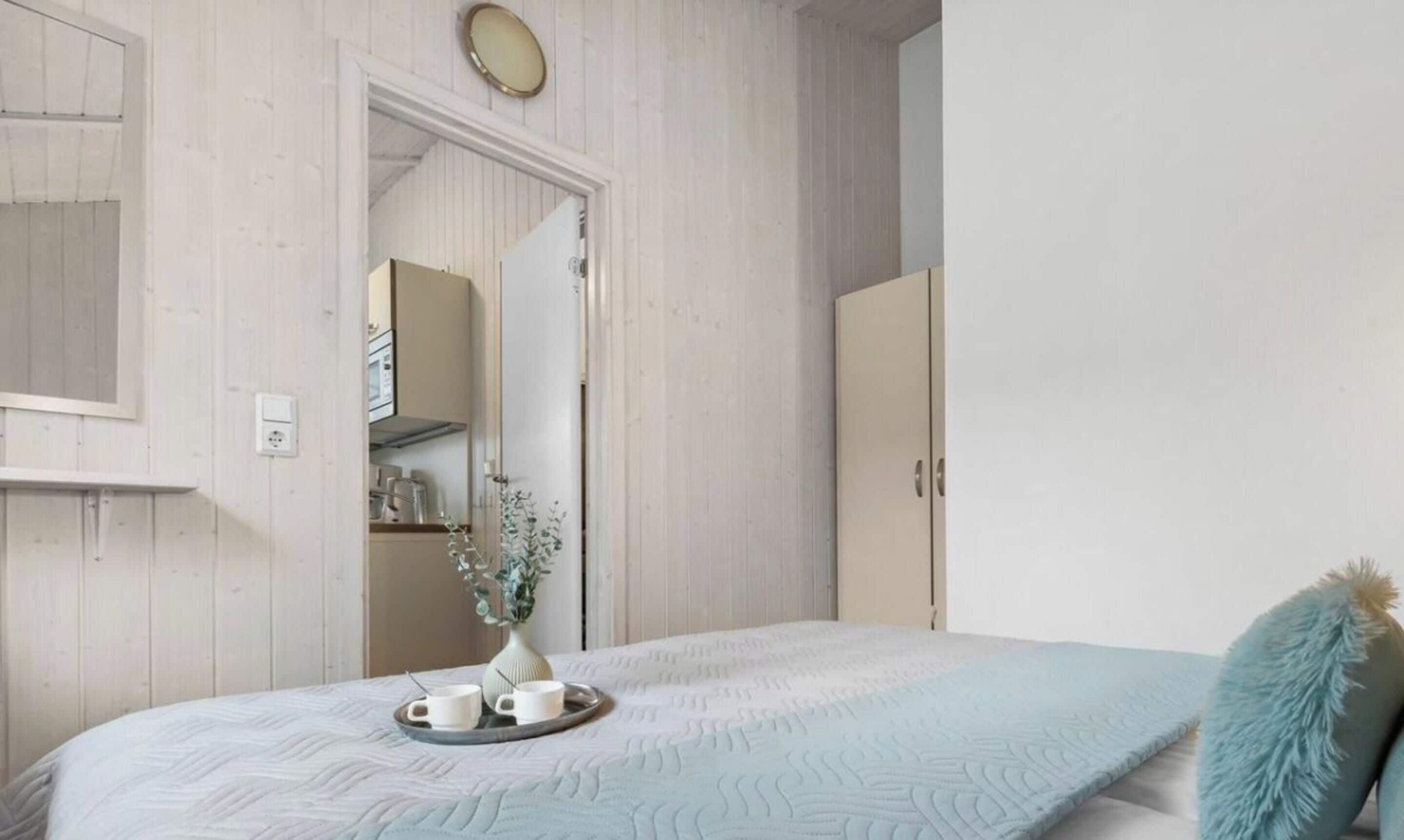 Un dormitorio con cama, una mesita de noche con tazas y bandeja, un armario y un espejo en la pared.
