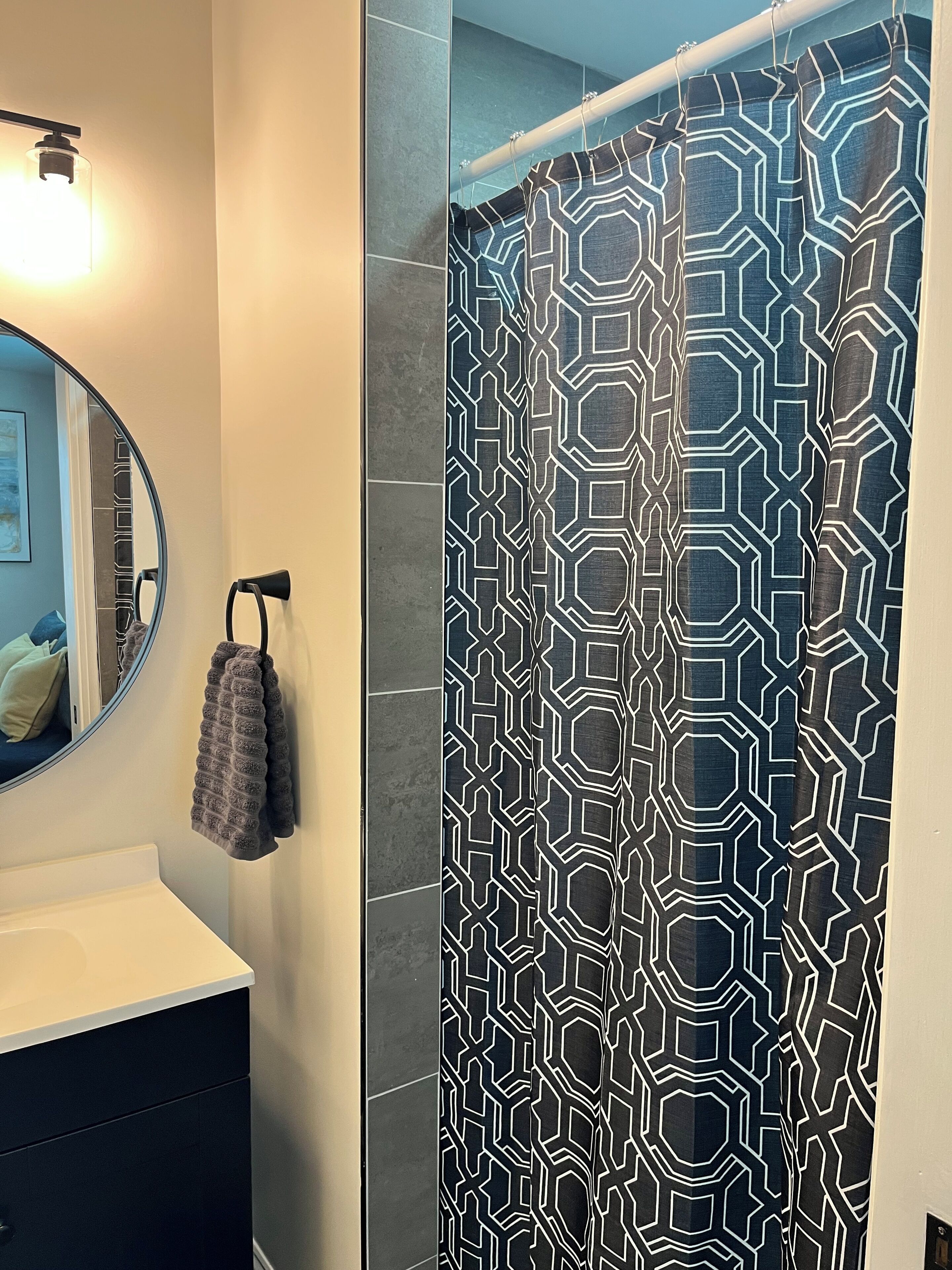 Ett badrum med modern design, med ett duschdraperi med geometriskt mönster, en rund spegel och en handdukshängare.