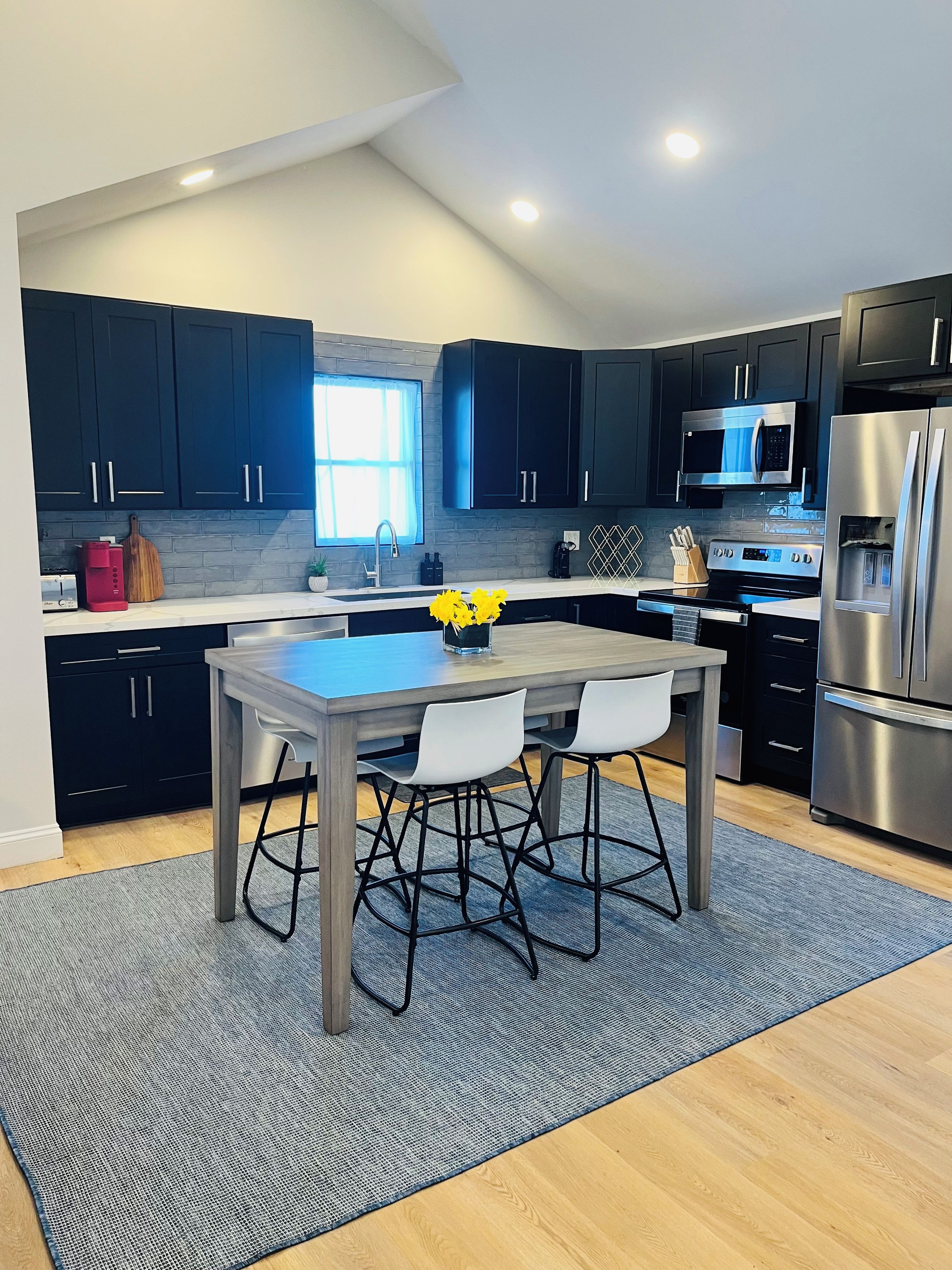 A modern kitchen with dark cabinets, a stainless steel refrigerator, and a dining area with a gray table and white chairs.