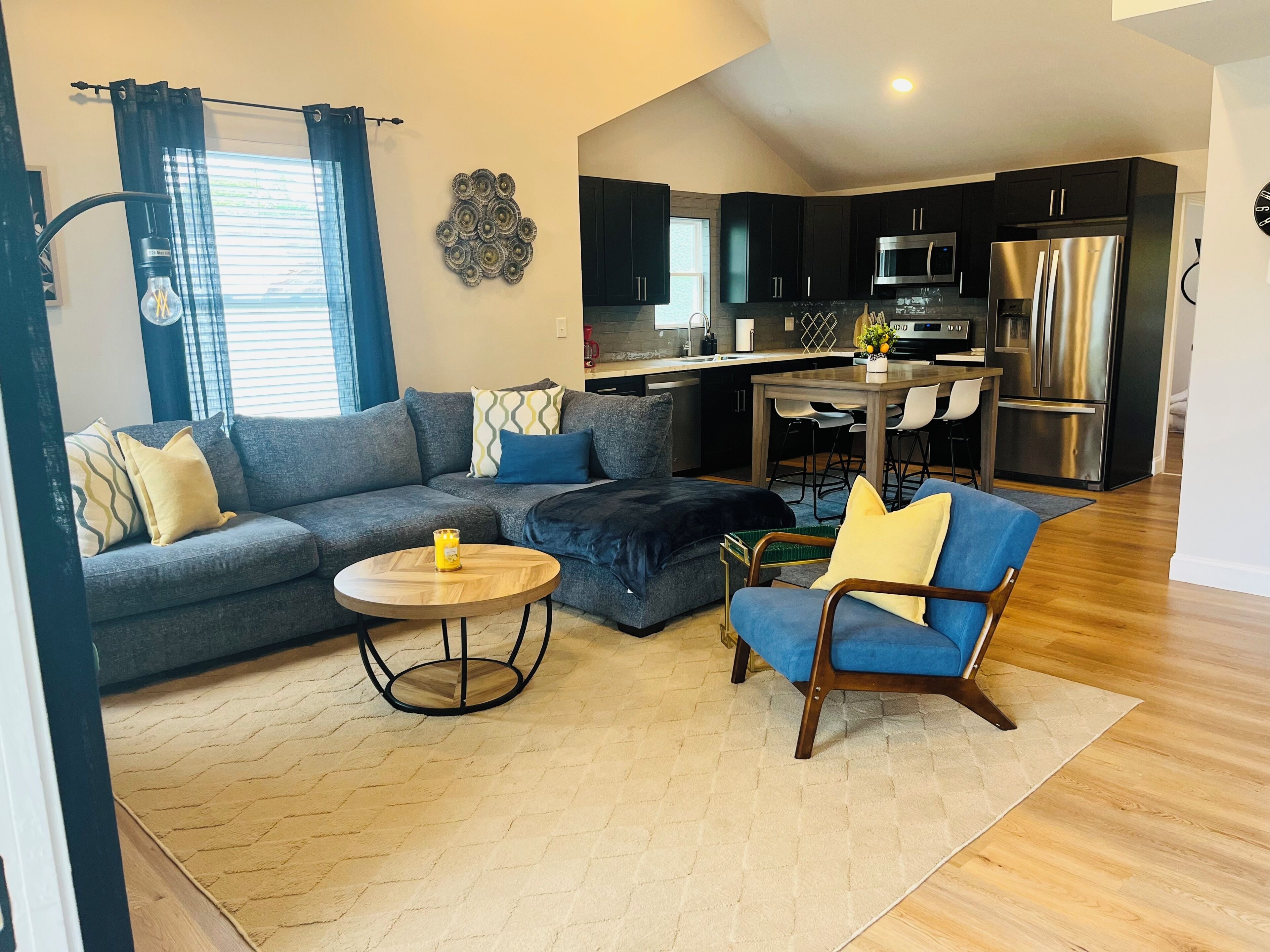A modern living room with a sofa, ottoman, and armchair, a dining area with a table and chairs, and a kitchen with stainless steel appliances.