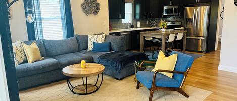 A modern living room with a sofa, ottoman, and armchair, a dining area with a table and chairs, and a kitchen with stainless steel appliances.
