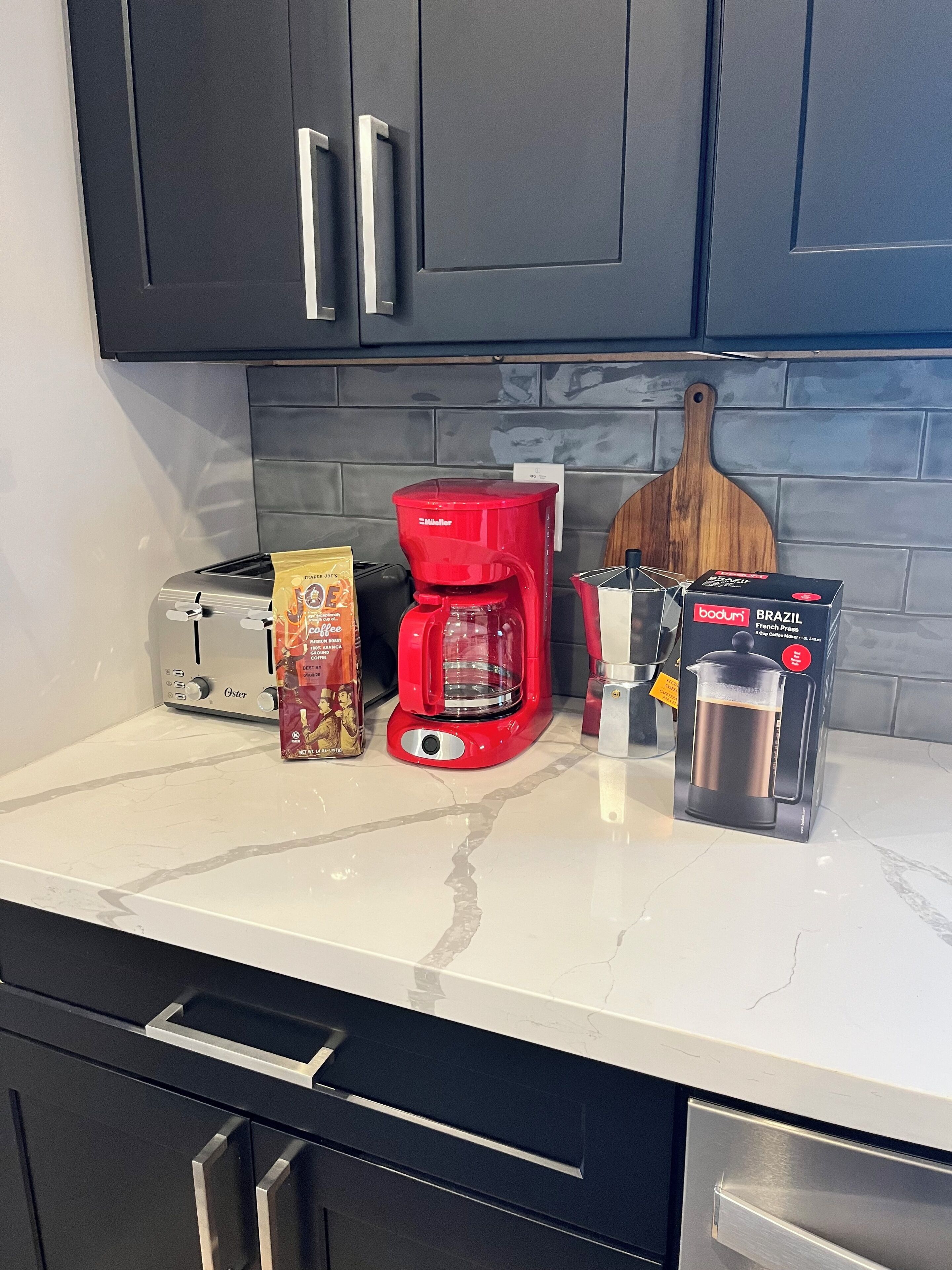 Una encimera de cocina con una cafetera roja, una prensa francesa, un tostador y un paquete de café.