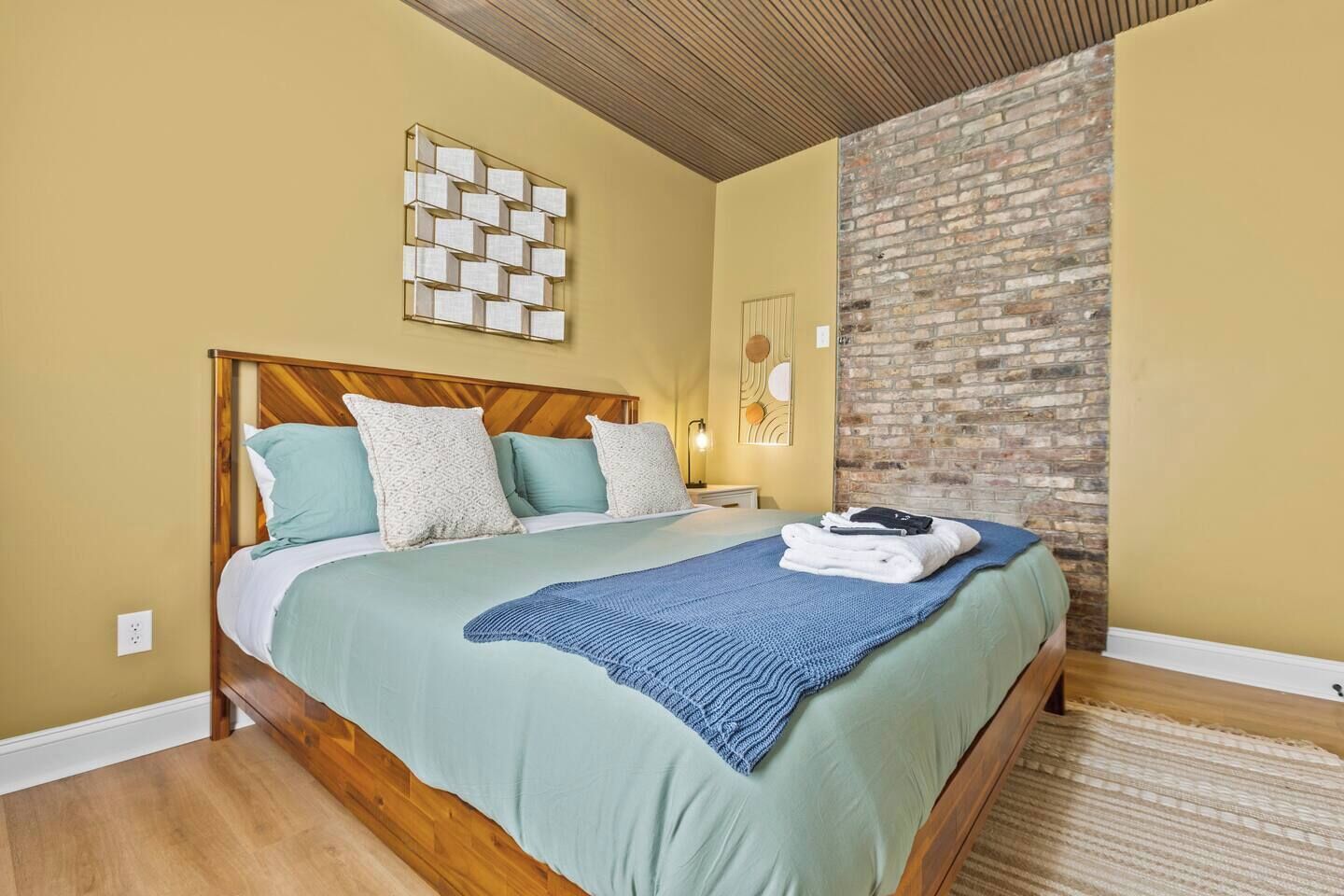 Una habitación de hotel con estructura de cama de madera, pared decorativa de ladrillo y una obra de arte moderna en la pared.