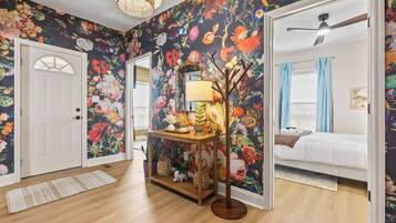 Una habitación con papel tapiz floral, piso de madera, un escritorio con una lámpara y una cama con cortina azul.