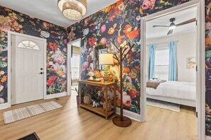 Habitació amb paper pintat floral, un paviment de fusta, una taula amb una làmpada i un llit amb cortina blava.