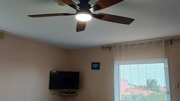 Une chambre dotée d’un ventilateur de plafond, d’une télévision, d’un lit recouvert d’une courtepointe bleue, d’une fenêtre ornée de rideaux en dentelle et d’une table sur laquelle se trouvent une lampe et des télécommandes.
