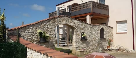 A two-story stone building with a balcony, a staircase, and a small garden area.