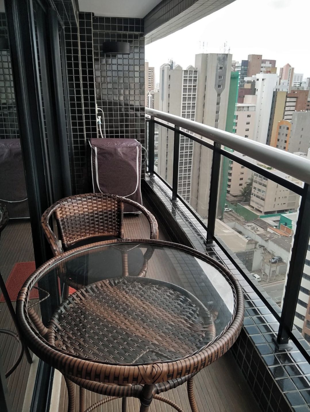 A balcony with wicker chairs and a glass table, overlooking a cityscape.