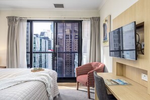 Ein Hotelzimmer mit Bett, Schreibtisch, Stuhl, Fernseher und einem großen Fenster mit Blick auf die Stadt.