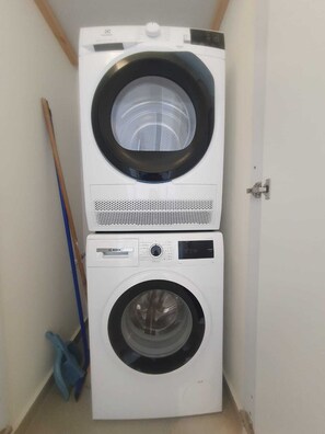 A stacked washer and dryer unit in a narrow space with a broom leaning against the wall. - Spacious New 1BD Balcony Mamad Levinski Florentin (Tel Aviv-Yafo)