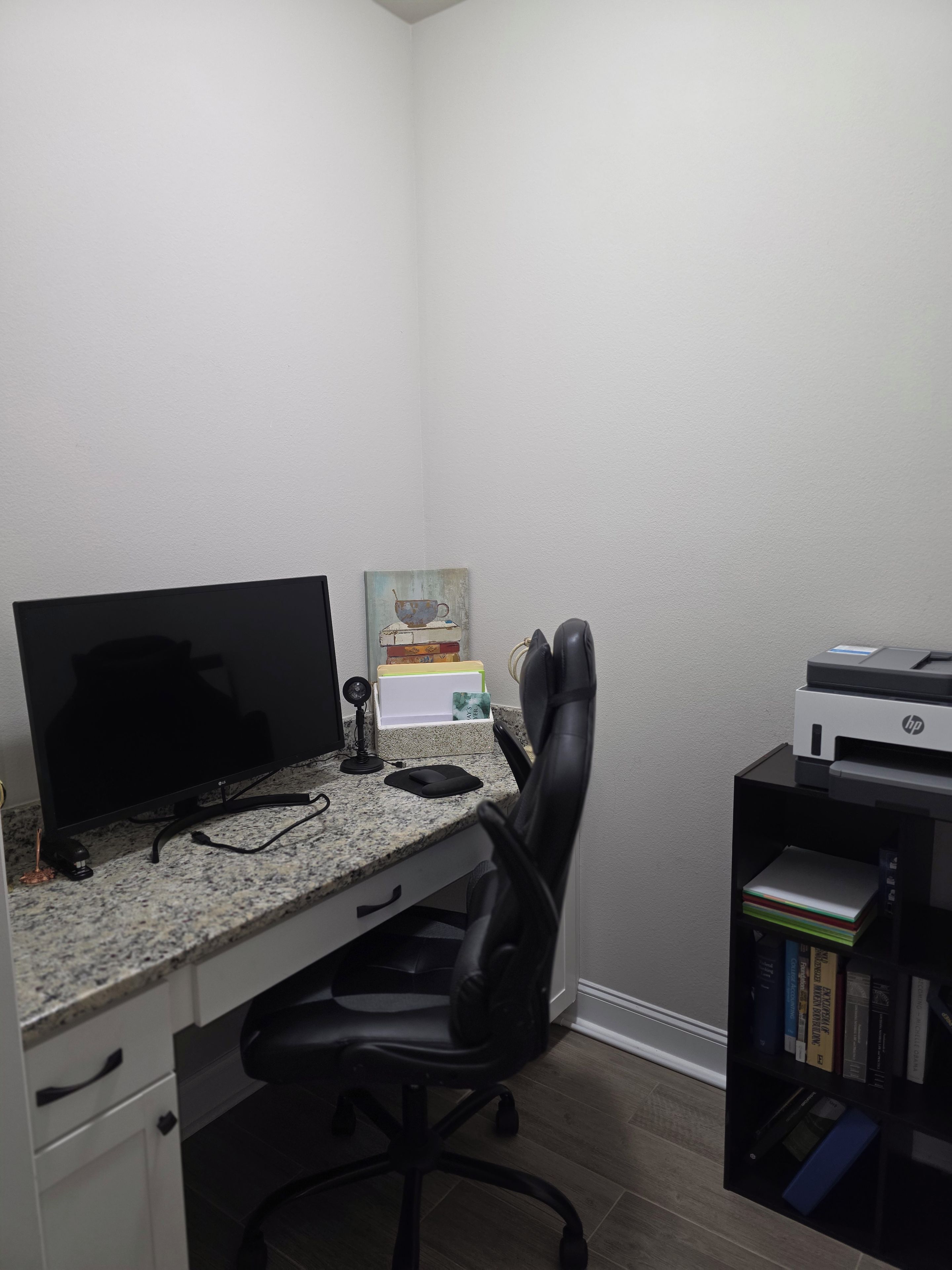 Ein Home-Office-Arbeitsplatz mit einem Schreibtischcomputer, einem schwarzen Bürostuhl, einem Drucker und einem Bücherregal.