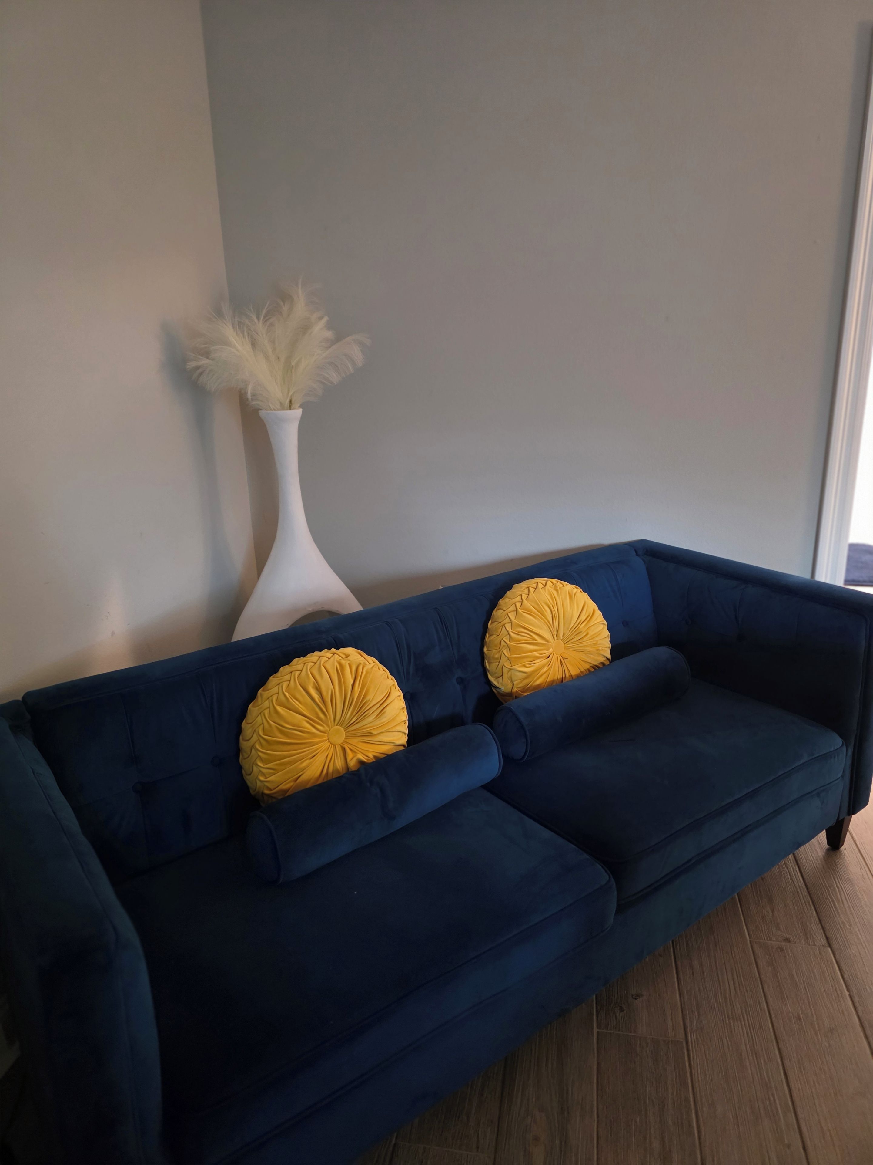 A dark blue sofa with two yellow pillows and a white vase with pampas grass against a plain wall.