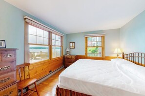 Quarto com cama grande, penteadeira de madeira e duas janelas com vista.