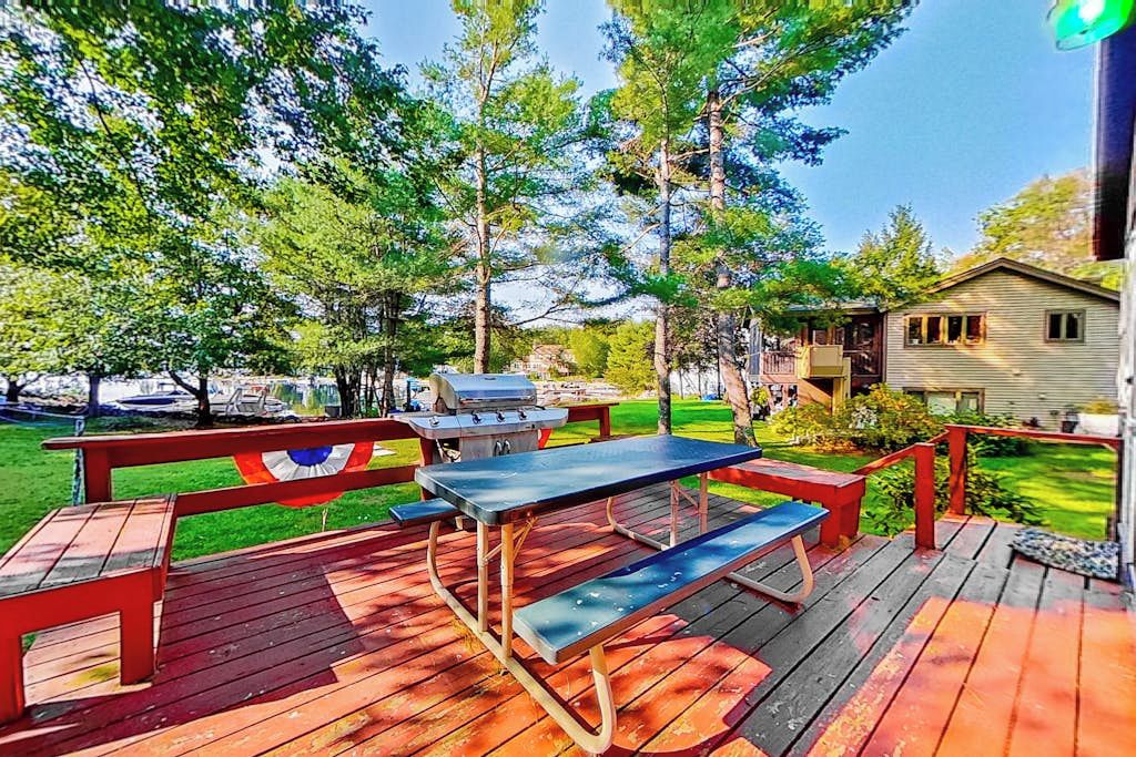 Una terraza de madera con mesas de picnic y una parrilla, rodeada de árboles y una casa.