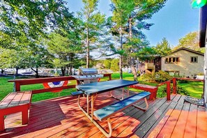 Eine Holzterrasse mit Picknicktischen und Grill, umgeben von Bäumen und einem Haus.