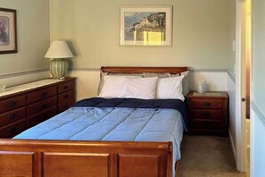 Quarto com cama de madeira, mesa de cabeceira, abajur, cômoda e um quadro na parede.