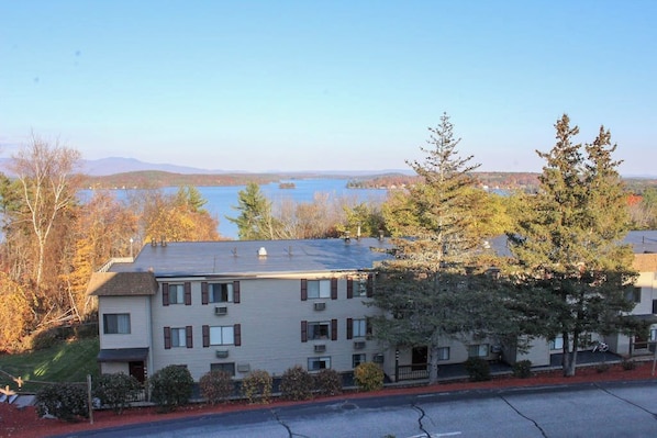 A multi-story building with a flat roof, surrounded by trees and a body of water in the background. - 3BR Near Golf - Balcony - Pool - Tennis Courts (Laconia)