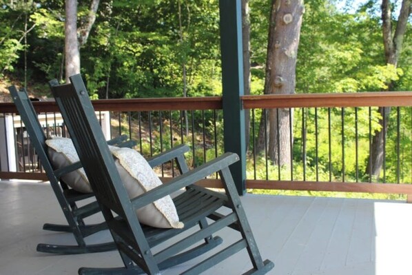 A rocking chair on a wooden deck with a railing, overlooking a forested area.