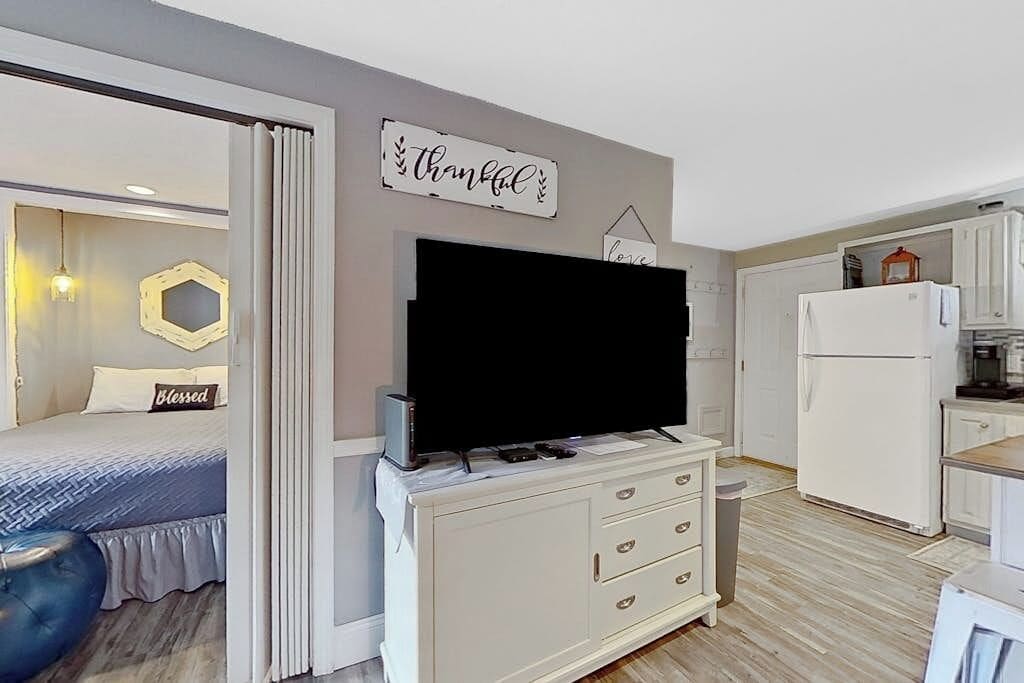A modern bedroom with a bed, a TV on a dresser, and a kitchen area with a refrigerator.
