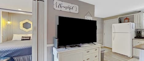 A modern bedroom with a bed, a TV on a dresser, and a kitchen area with a refrigerator.