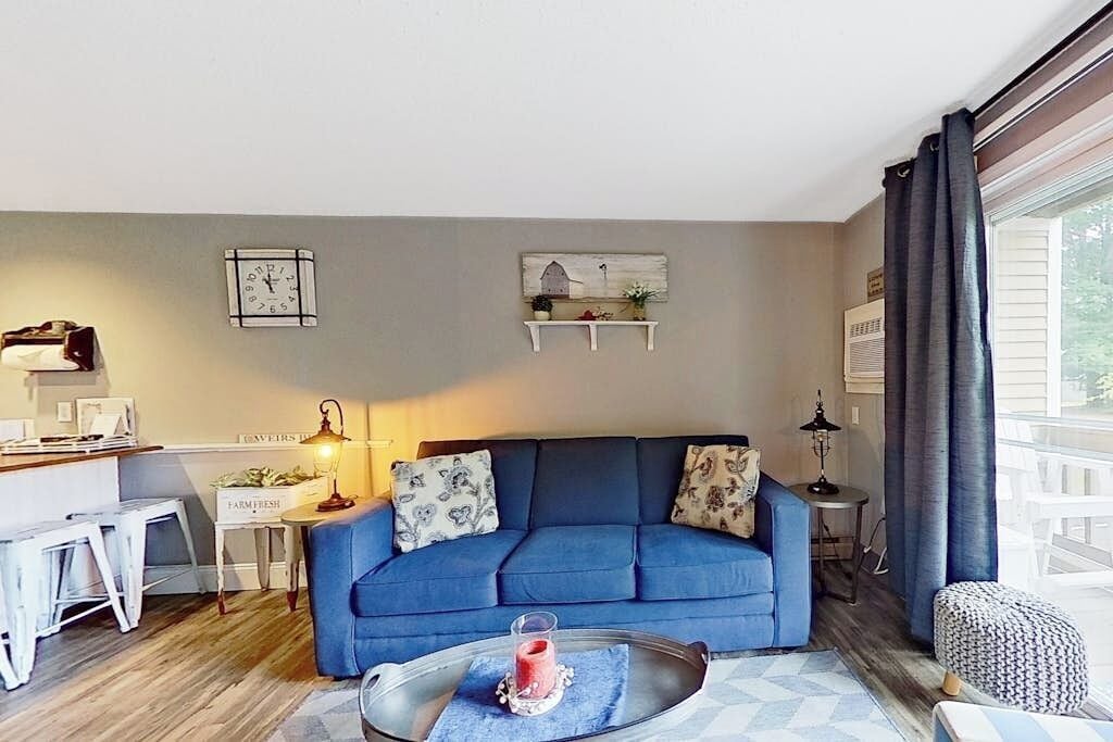 A living room with a blue sofa, a coffee table with a candle, and a kitchen area with stools.