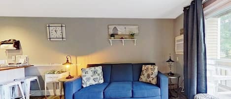 A living room with a blue sofa, a coffee table with a candle, and a kitchen area with stools.
