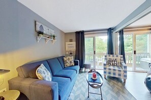 A living room with a blue sofa, a small table with a candle, a chair with a cushion, and a balcony with a view. - 1BR with beach and lake access, pool, & sports courts (Gilford)
