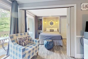A bedroom with a bed, a chair with a cushion, a round ottoman, and a view of the outdoors.
