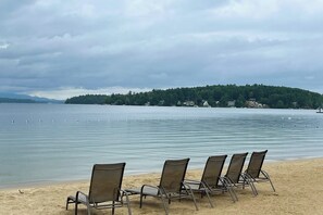 A beach with four metal chairs lined up facing the water, a calm lake, and a forested hill in the background. - 1BR with beach and lake access, pool, & sports courts (Gilford)