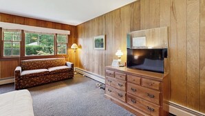 A hotel room with a wooden paneled wall, a sofa, a TV on a wooden cabinet, and a window with a view of greenery.