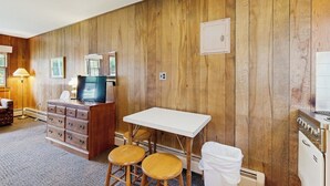 A room with wooden panel walls, a white table with two stools, a wooden dresser, a television, and a white radiator.