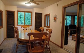 A dining room with a large wooden table and chairs, a ceiling fan, and windows with shutters.