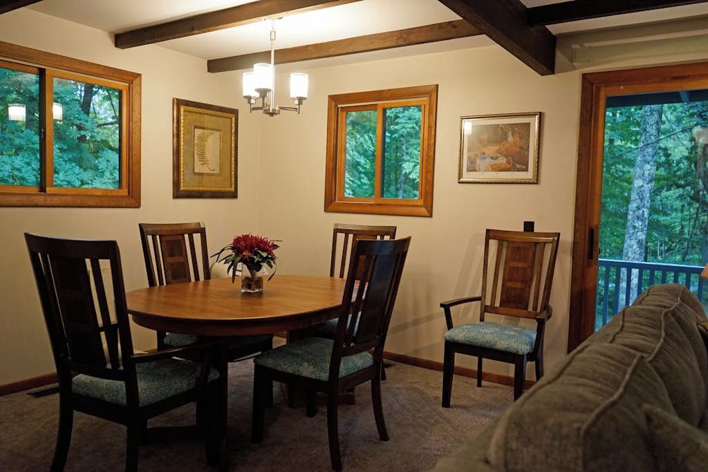 A dining area with a round table, six chairs, and a vase with flowers. There are framed pictures on the walls and windows with a view of trees.