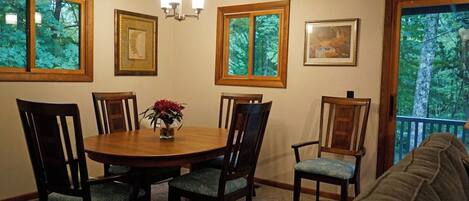 A dining area with a round table, six chairs, and a vase with flowers. There are framed pictures on the walls and windows with a view of trees.