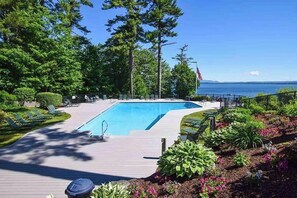 A swimming pool with a deck, surrounded by greenery and a view of the water. - Lake-view 2BR with lakefront swimming, pools (Gilford)