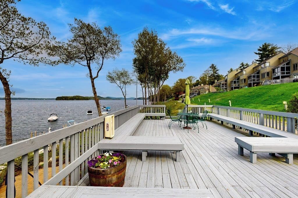 A wooden deck with benches, overlooking a body of water with boats, and a row of buildings in the background.