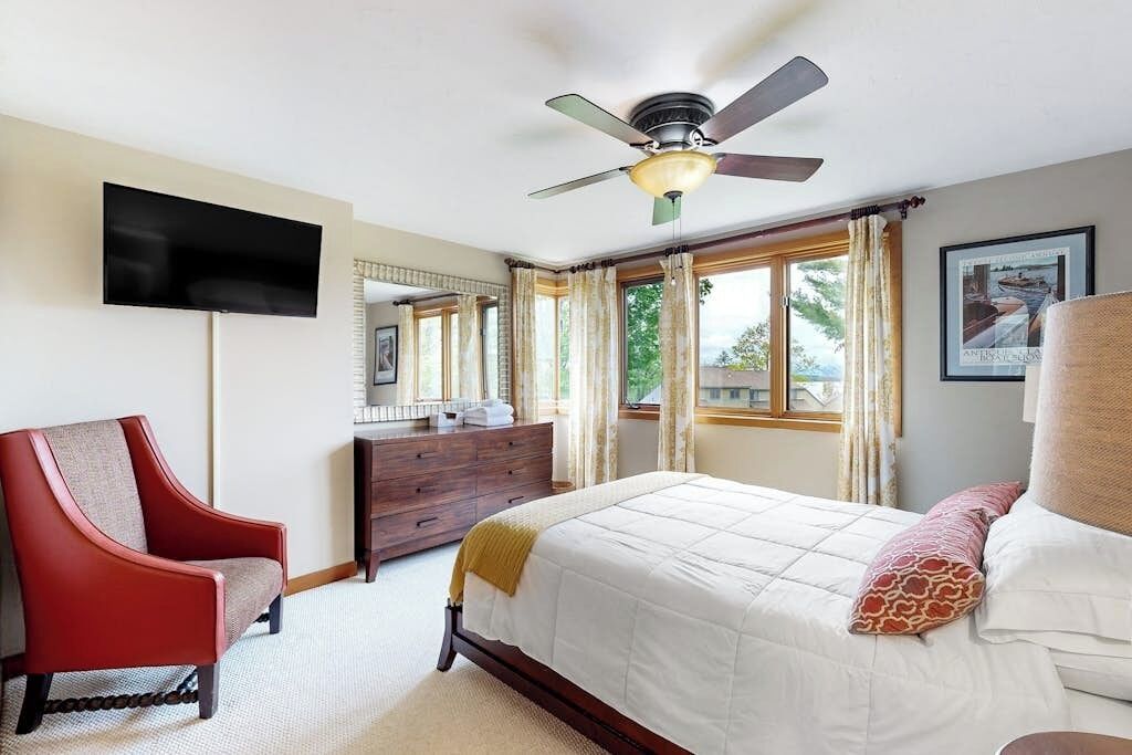 A hotel room with a bed, a red armchair, a dresser, a flat-screen TV, and a ceiling fan.