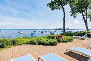 A waterfront patio with lounge chairs, a brick pathway, and boats on the water. - Lake-view 2BR with lakefront swimming, pools (Gilford)
