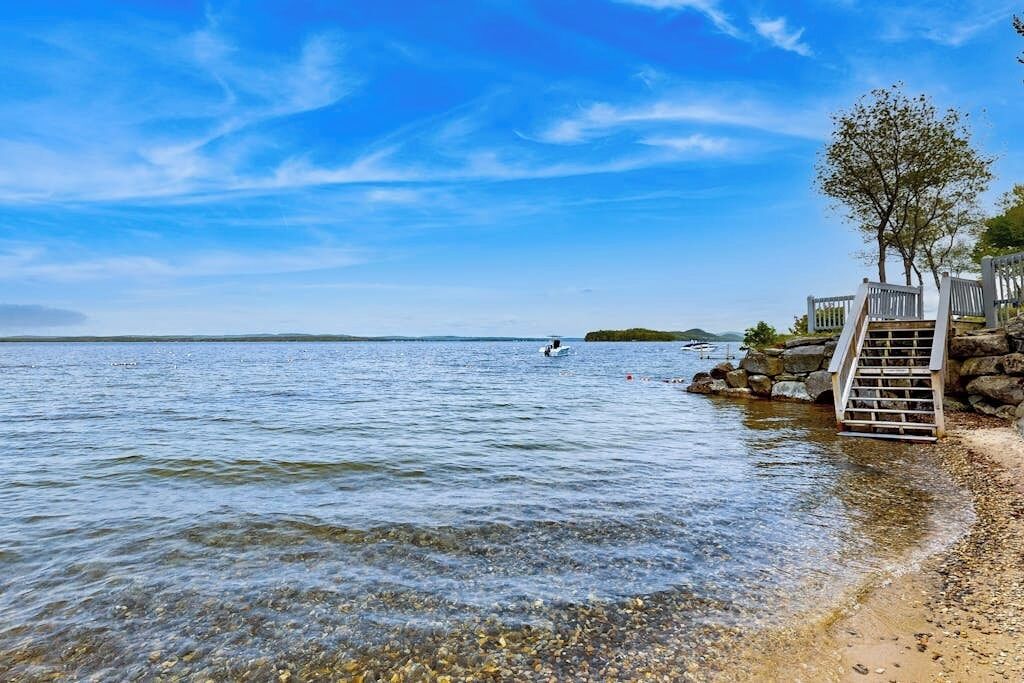 A clear lake with a rocky shore, a wooden staircase leading down to the water, and a tree on the right.