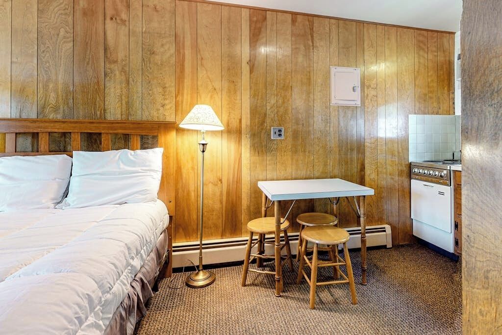 Una camera d'albergo con un letto, un tavolo con due sedie, una lampada e un angolo cottura.